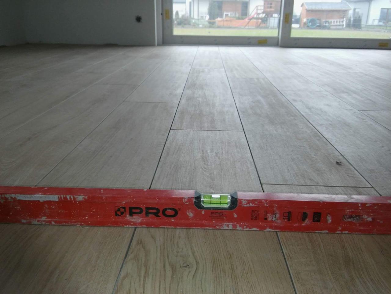 Ułożone płytki podłogowe imitujące drewno, wyrównywane czerwoną poziomicą PRO, widok na okno i dom w tle.