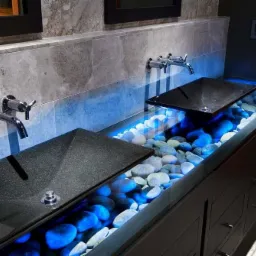 Nowoczesna łazienka z dwoma czarnymi umywalkami nablatowymi, podświetlanymi niebieskim światłem kamieniami dekoracyjnymi i chromowaną armaturą zamontowaną na szarej, marmurkowej ścianie.