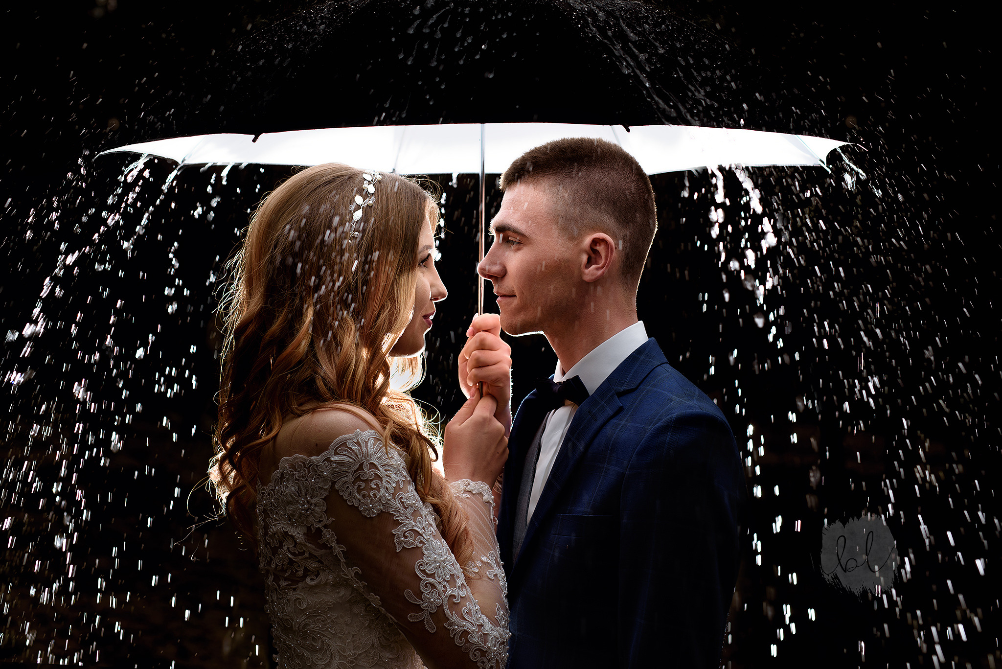 Para młoda, panna młoda w koronkowej sukni i pan młody w granatowym garniturze pod białym parasolem w strugach deszczu, profil, ciemne tło.