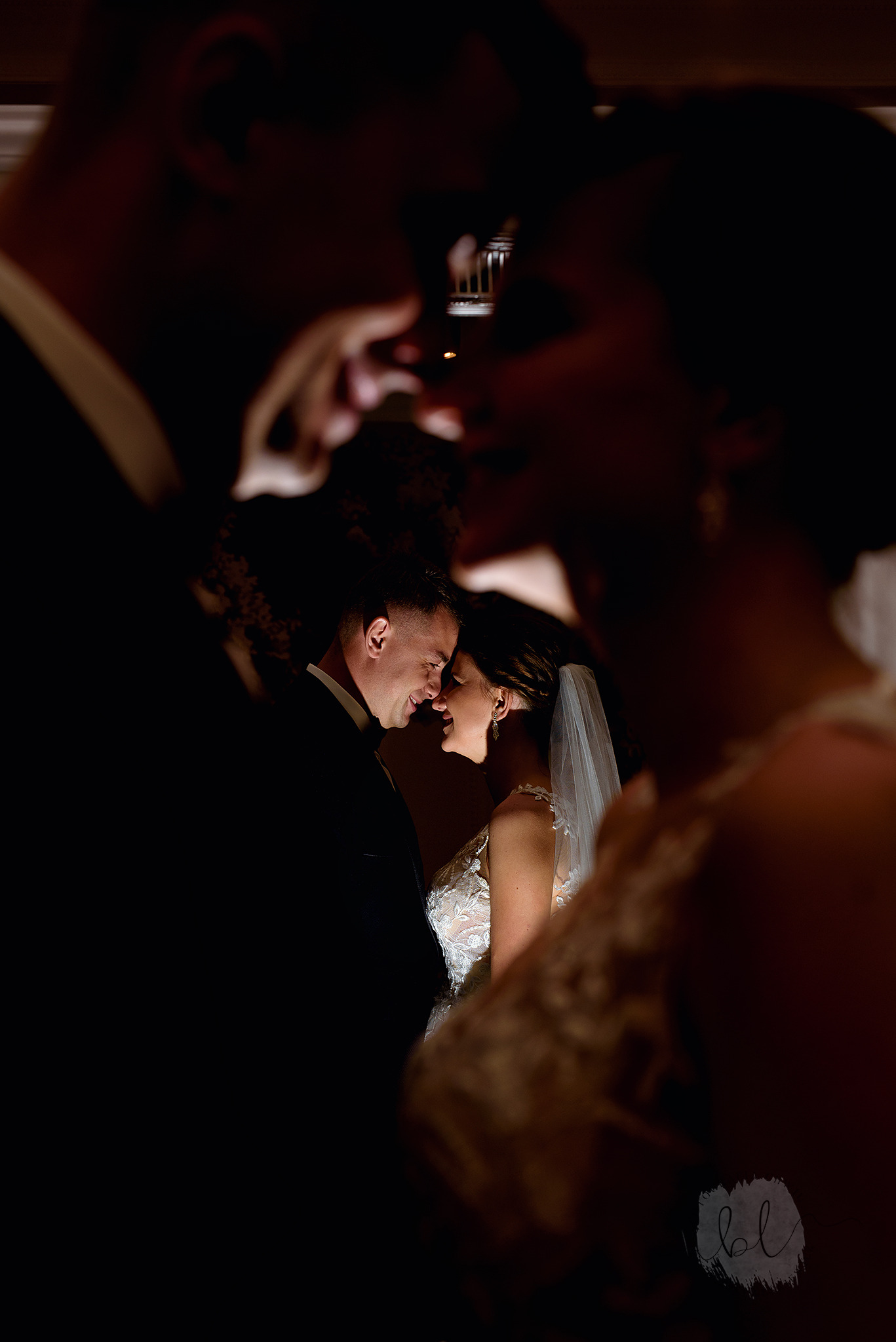 Nowożeńcy delikatnie dotykają się nosami, panna młoda w koronkowej sukni i welonie, pan młody w czarnym smokingu, ujęcie zza ramion gości weselnych.