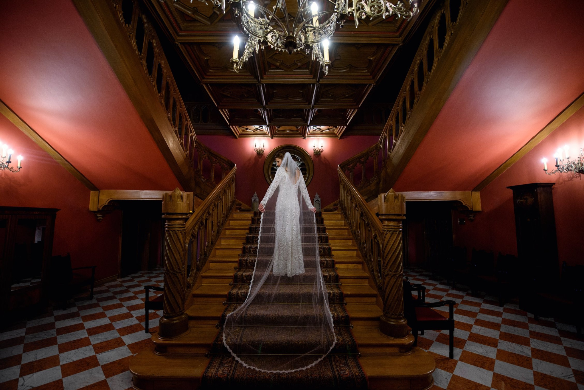 Panna młoda w białej sukni, z długim welonem, pozuje na drewnianych schodach w zabytkowym wnętrzu z czerwonymi ścianami i wzorzystą podłogą.