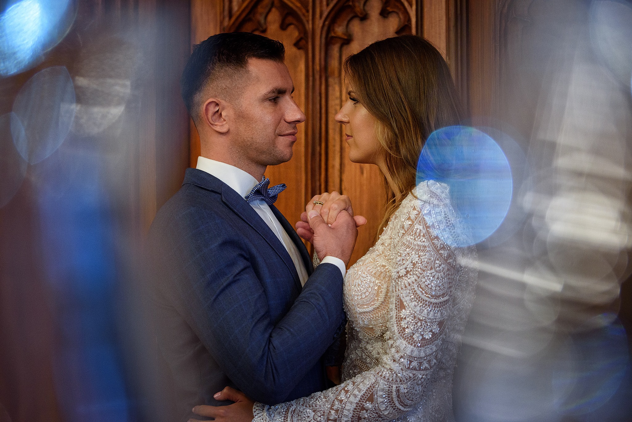 Para młoda trzymająca się za ręce, on w granatowym garniturze z muszką, ona w koronkowej sukni, oboje patrzą na siebie na tle rzeźbionych drewnianych drzwi, z efektem bokeh