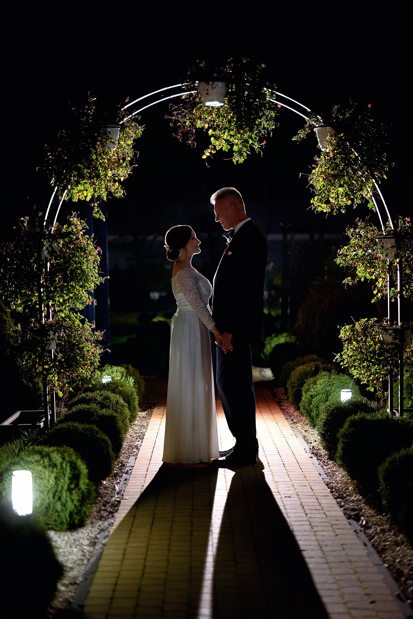 Para młoda trzymająca się za ręce pod oświetlonym, kwiatowym łukiem w nocy, z długimi cieniami na brukowanej ścieżce.