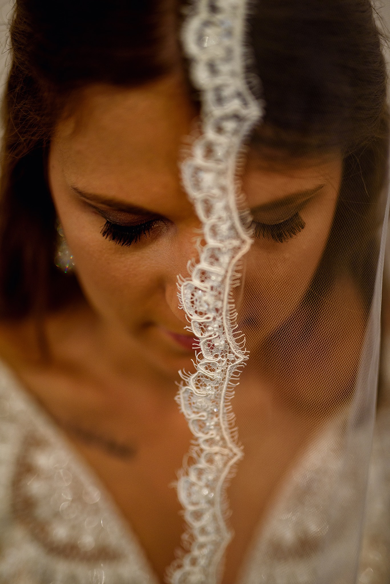 Panna młoda z długimi rzęsami, spoglądająca w dół, częściowo zakryta delikatnym, koronkowym welonem z połyskującymi zdobieniami, w tle fragment sukni ślubnej.