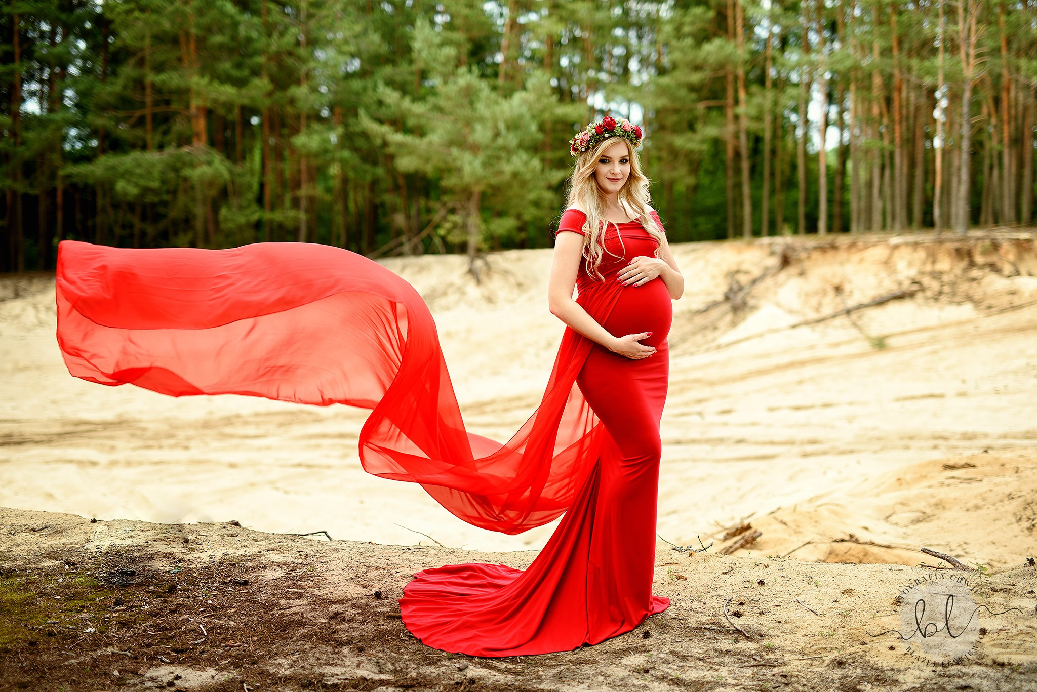 Kobieta w czerwonej sukni i kwiatowym wianku, trzymająca się za brzuch, na tle piaszczystego krajobrazu i lasu iglastego, z powiewającą na wietrze czerwoną tkaniną.