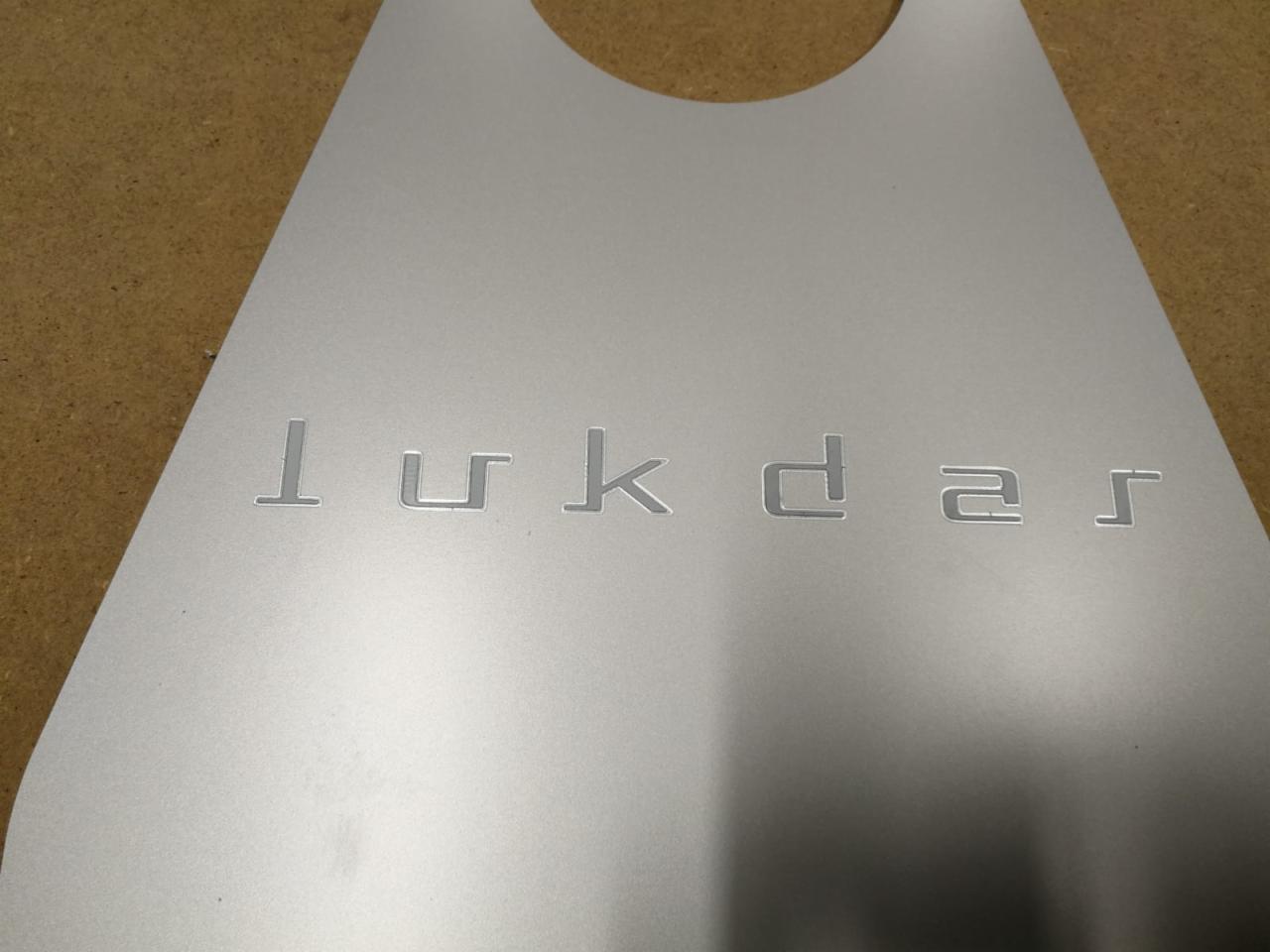 Szary, metalowy element z wygrawerowanym napisem 'lukdar' w nowoczesnej czcionce, o zaokrąglonym kształcie u góry, leżący na brązowym tle.