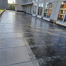 Roof professional Łukasz Bartkowski - Nowa, czarna, płaska powierzchnia dachu z papy bitumicznej na budynku w trakcie budowy, widoczne odciski butów i odbicia w mokrej powierzchni.