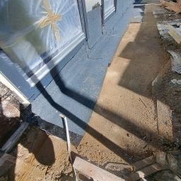 Roof professional Łukasz Bartkowski - Przygotowanie elewacji budynku z oknami zabezpieczonymi folią, widoczna warstwa izolacyjna i grunt na fragmencie ściany, teren wokół w trakcie prac budowlanych.