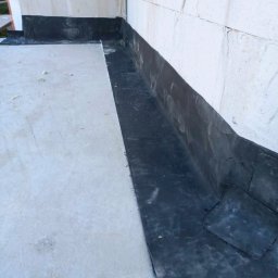 Roof professional Łukasz Bartkowski - Hydroizolacja w narożniku dachu płaskiego wykonana z czarnej papy bitumicznej, widoczna ściana z bloczków betonowych.
