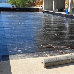 Roof professional Łukasz Bartkowski - Hydroizolacja płaskiego dachu papą termozgrzewalną, widoczna butla z gazem i palnik, w tle wieża ciśnień i jezioro.