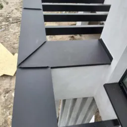 Szczegółowe ujęcie nowej, czarnej, metalowej obróbki blacharskiej dachu, z widocznymi łączeniami i fragmentem elewacji w surowym tynku.