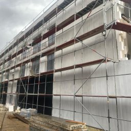 Roof professional Łukasz Bartkowski - Budynek w trakcie ocieplania elewacji, widoczne rusztowania, styropianowe płyty dociepleniowe i częściowo nałożony tynk, przygotowane materiały budowlane na ziemi.