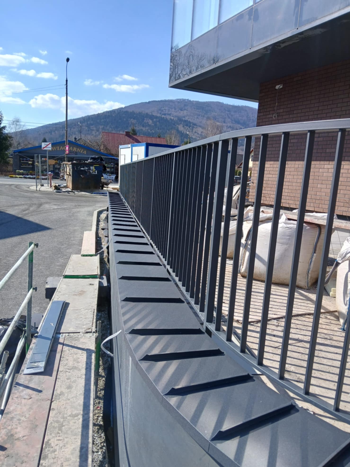 Nowoczesna elewacja budynku w trakcie wykończenia z widocznymi rusztowaniami i workami z materiałami budowlanymi w tle.