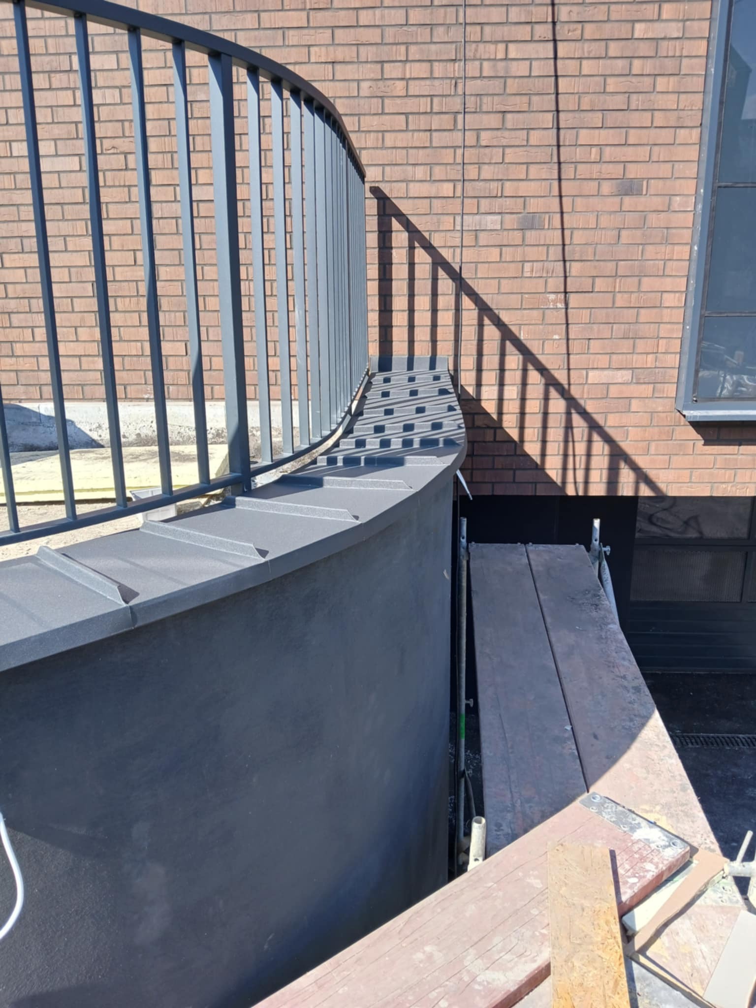 Szczegółowe ujęcie obróbki blacharskiej dachu z balustradą w Rzozowie, widoczne rusztowanie i cień na ceglanej ścianie.