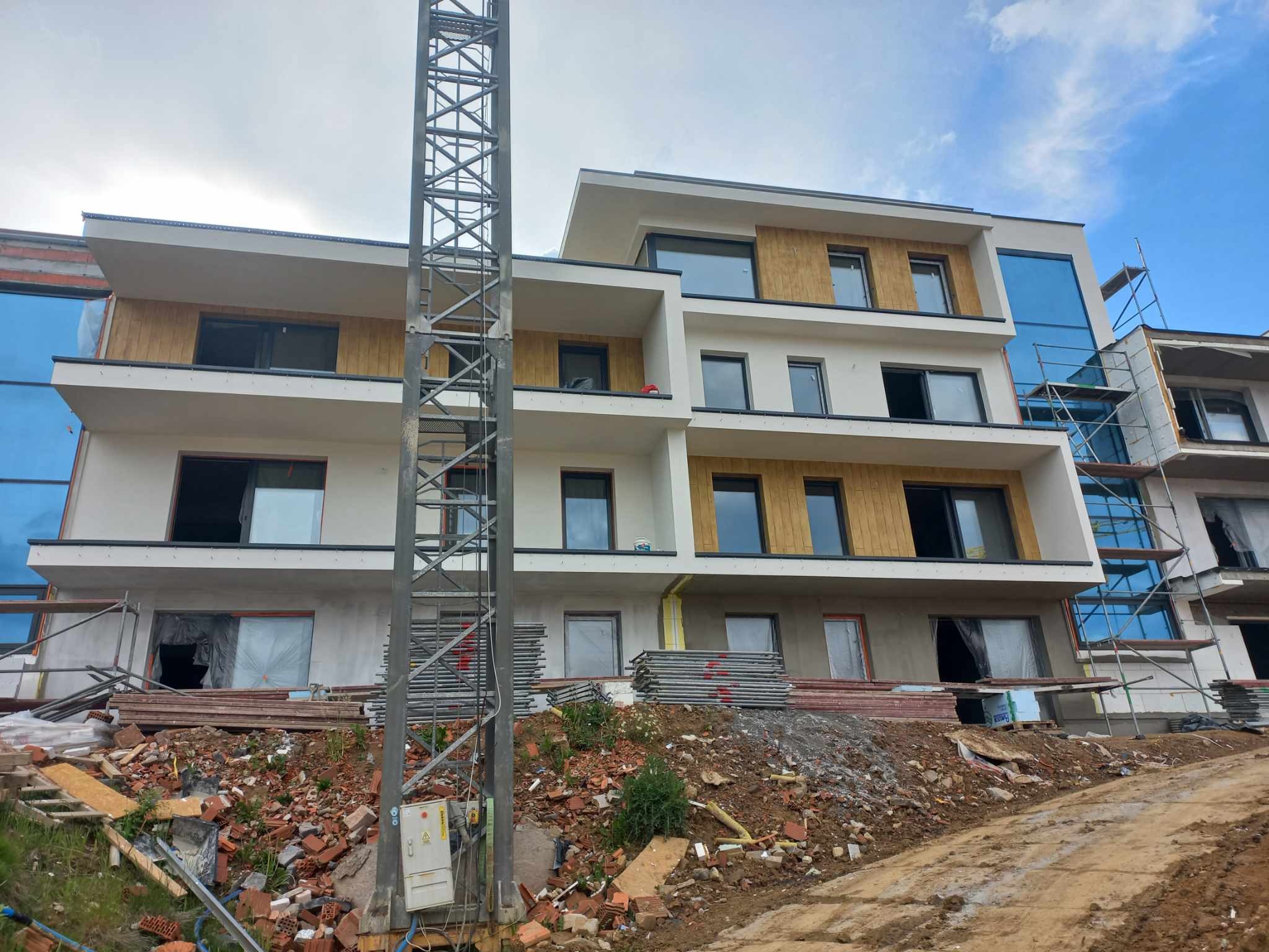 Wielopiętrowy budynek mieszkalny w trakcie prac elewacyjnych, z widocznymi rusztowaniami, stertą materiałów budowlanych i metalową konstrukcją na pierwszym planie.