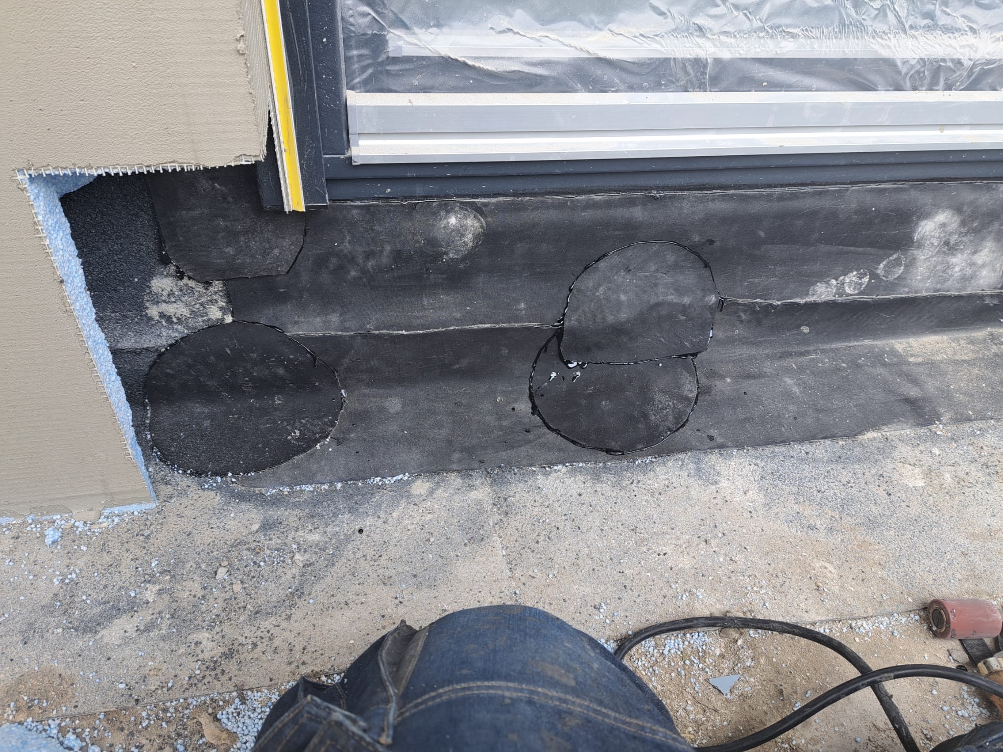 Fragment dachu płaskiego z papy bitumicznej z okrągłymi wycięciami, widoczna warstwa ocieplenia elewacji i fragment okna dachowego, na podłożu rozsypane drobne elementy.