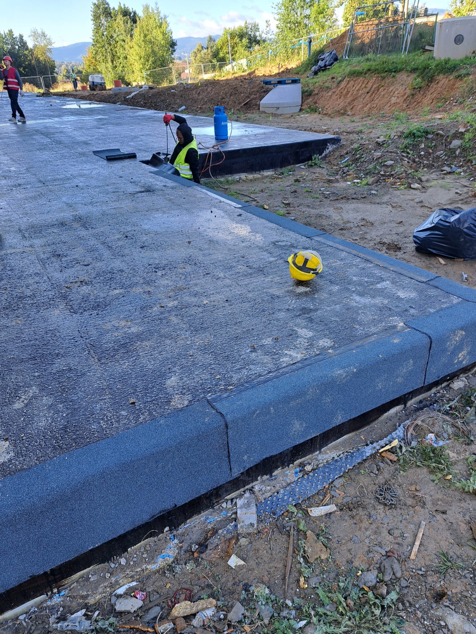 Praca dekarza przy izolacji płaskiego dachu papą termozgrzewalną, widoczna butla z gazem, palnik, kask ochronny i obrzeże dachu z kostki. W tle krajobraz z drzewami i zabudowaniami.