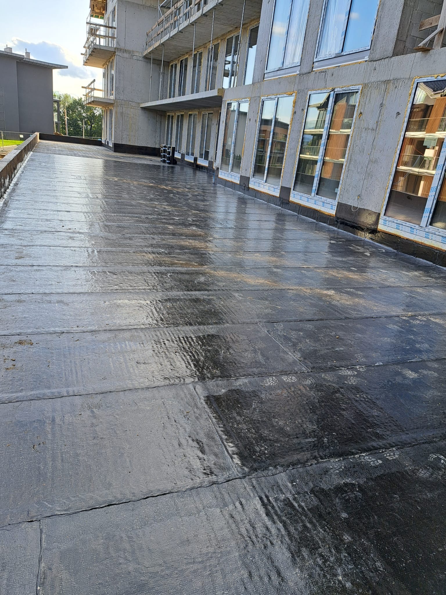 Nowa, czarna, płaska powierzchnia dachu z papy bitumicznej na budynku w trakcie budowy, widoczne odciski butów i odbicia w mokrej powierzchni.