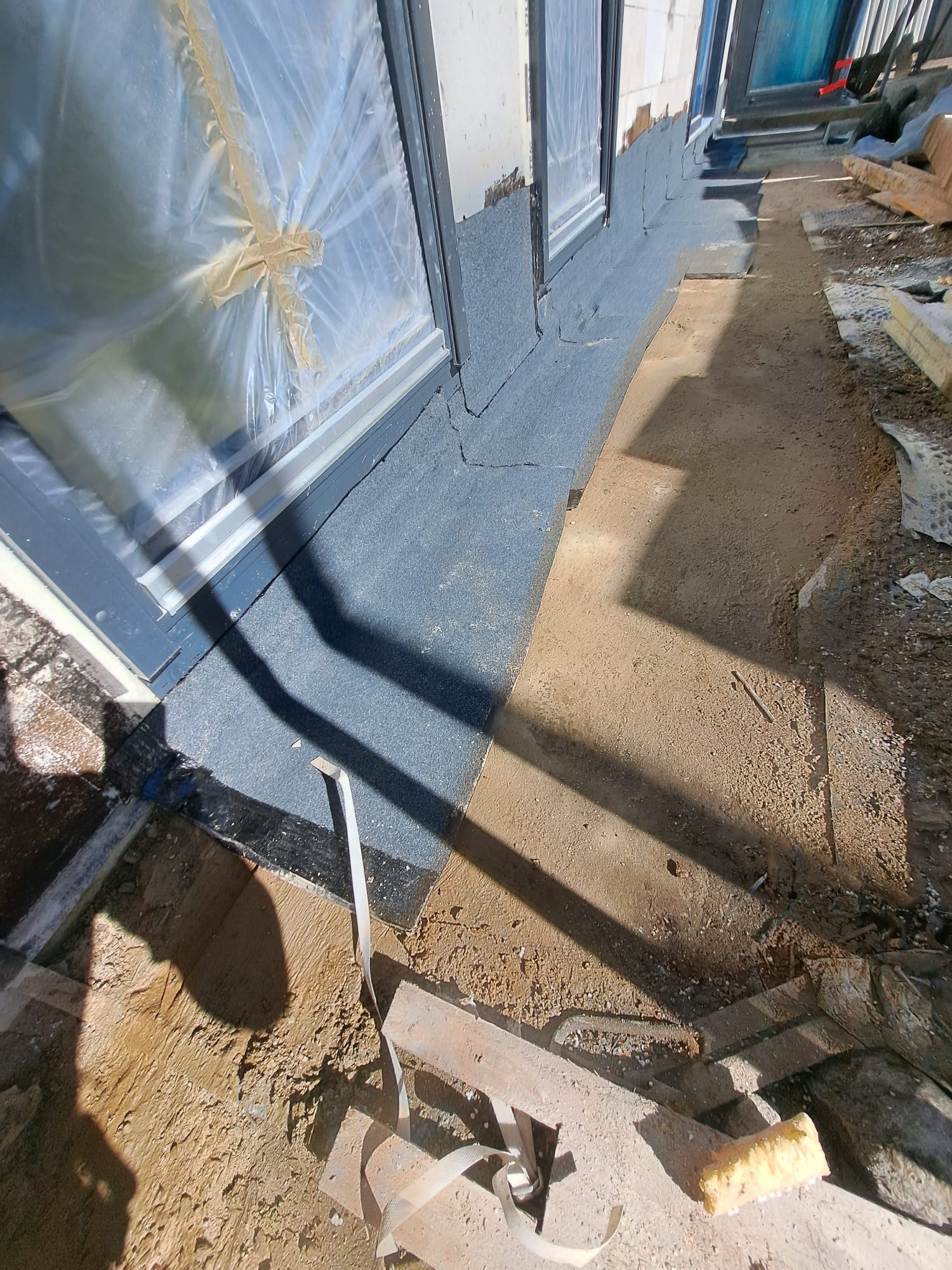 Przygotowanie elewacji budynku z oknami zabezpieczonymi folią, widoczna warstwa izolacyjna i grunt na fragmencie ściany, teren wokół w trakcie prac budowlanych.