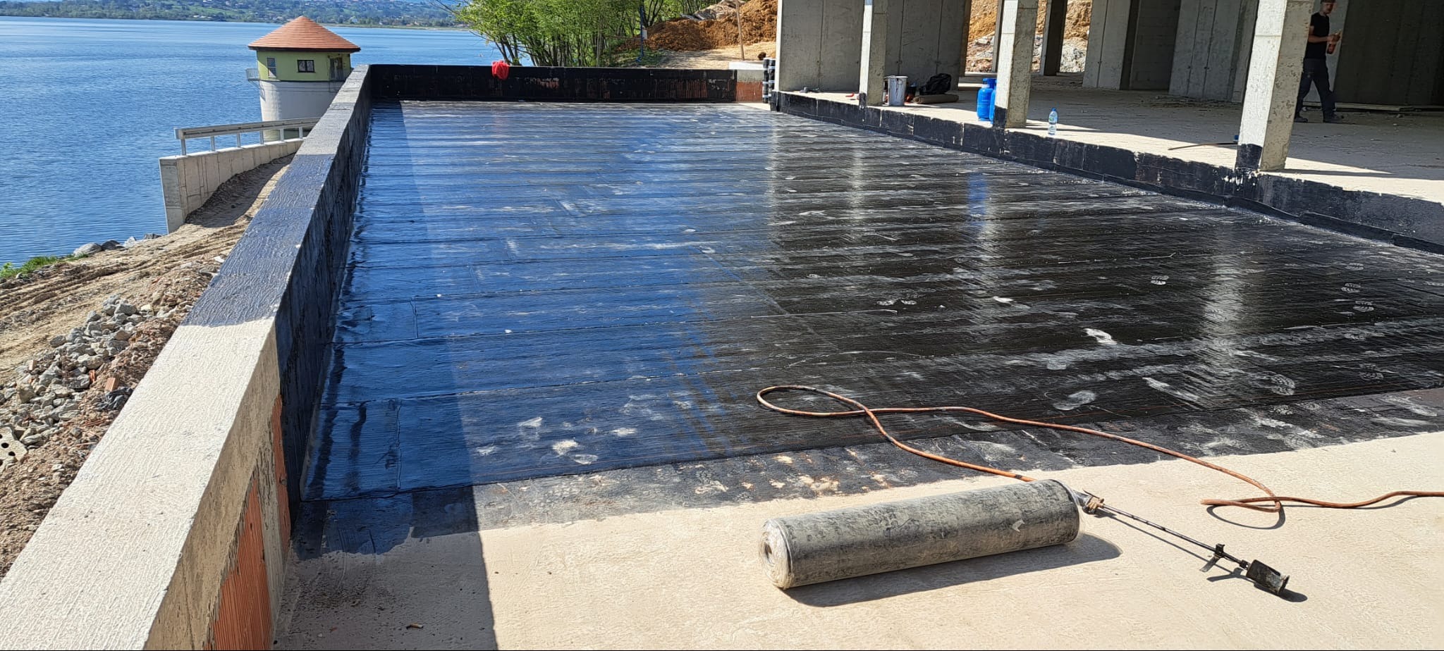 Hydroizolacja płaskiego dachu papą termozgrzewalną, widoczna butla z gazem i palnik, w tle wieża ciśnień i jezioro.