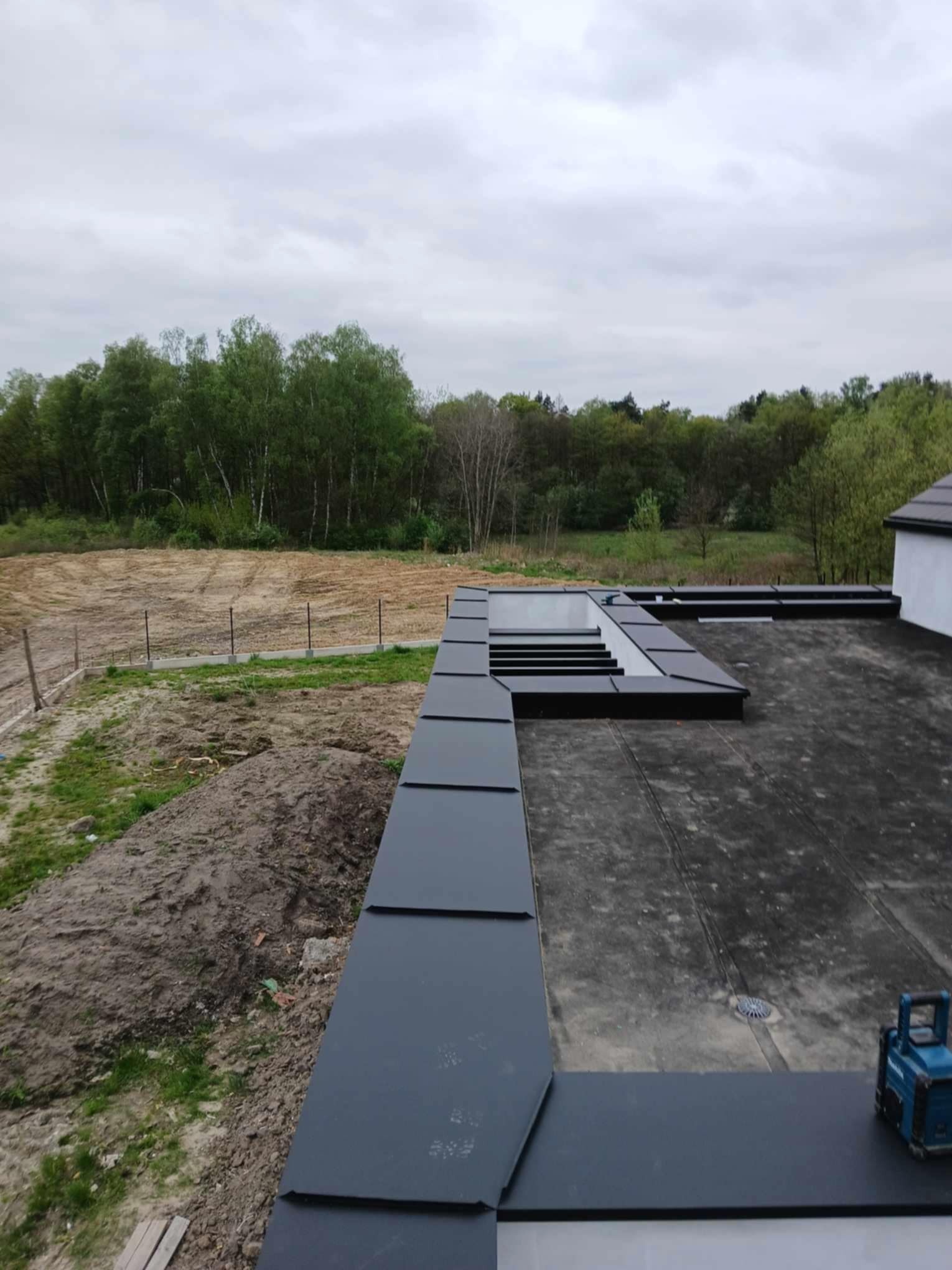 Widok z góry na płaski dach z czarną blachą i świetlikami dachowymi, na tle zieleni drzew i przygotowanego terenu budowlanego, z widocznym narzędziem dekarskim.