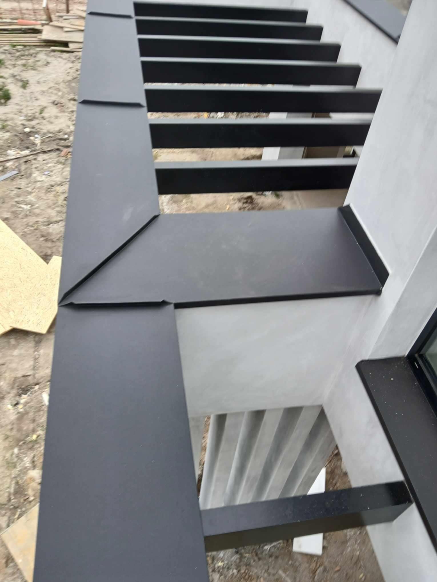 Szczegółowe ujęcie nowej, czarnej, metalowej obróbki blacharskiej dachu, z widocznymi łączeniami i fragmentem elewacji w surowym tynku.