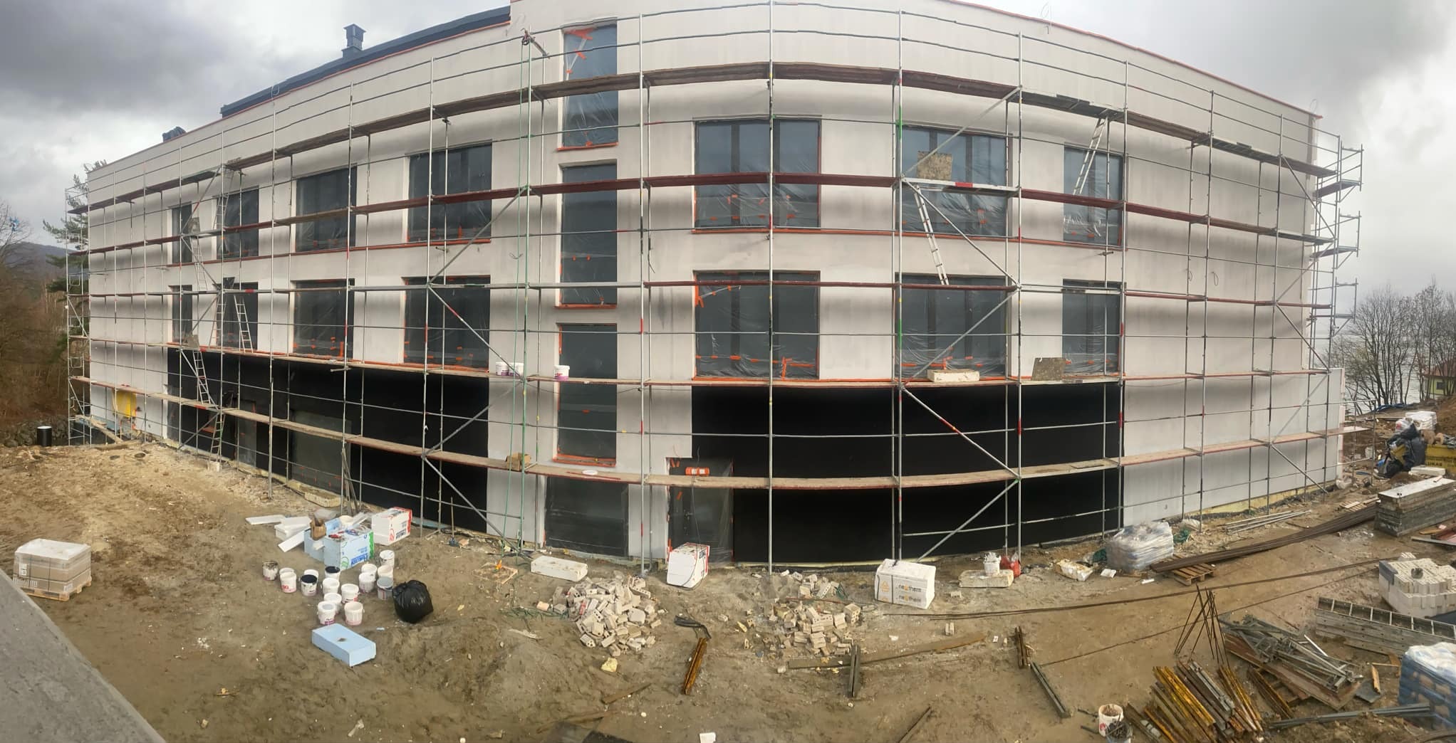 Budynek w trakcie ocieplania elewacji, widoczne rusztowania, okna zabezpieczone folią, przygotowane materiały budowlane i farby.