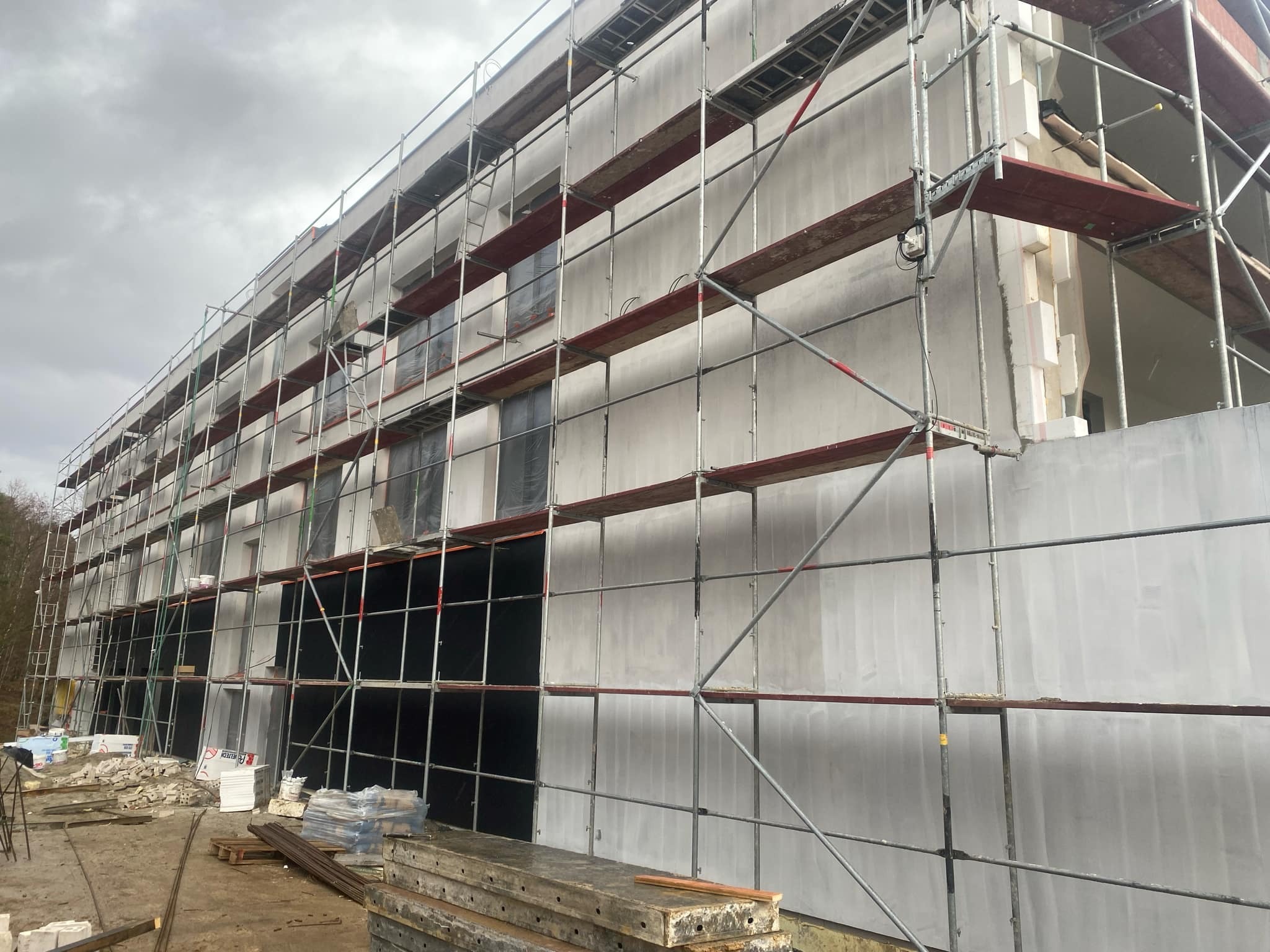 Budynek w trakcie ocieplania elewacji, widoczne rusztowania, styropianowe płyty dociepleniowe i częściowo nałożony tynk, przygotowane materiały budowlane na ziemi.