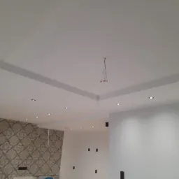 Biały sufit podwieszany z wpuszczonymi punktowymi lampami LED, fragment ściany z dekoracyjną tapetą w tle, widoczne przewody elektryczne.