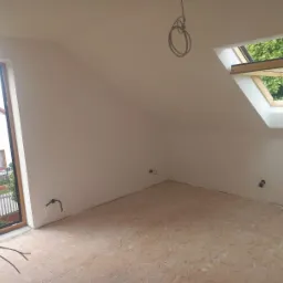 Poddasze po malowaniu ścian na biało, widoczne okno dachowe z widokiem na zieleń, surowa podłoga z płyt OSB, wystające kable elektryczne.