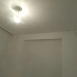 Wnętrze w Jelnej w trakcie prac wykończeniowych, widoczne gładkie, białe ściany przygotowane pod malowanie, surowa żarówka zwisająca z sufitu i wystające kable.