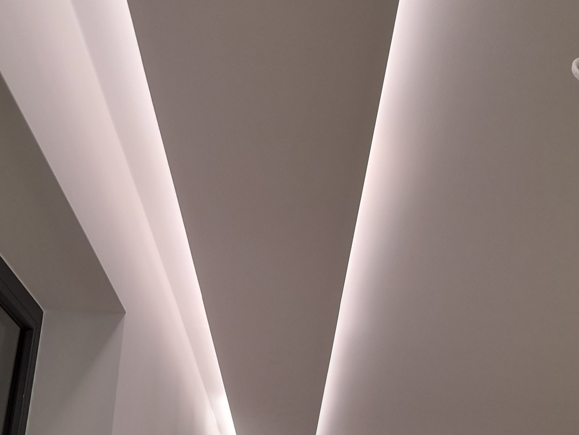 Nowoczesny sufit z wbudowanym oświetleniem LED tworzącym geometryczne wzory, widoczny fragment ramy okiennej w lewym dolnym rogu.