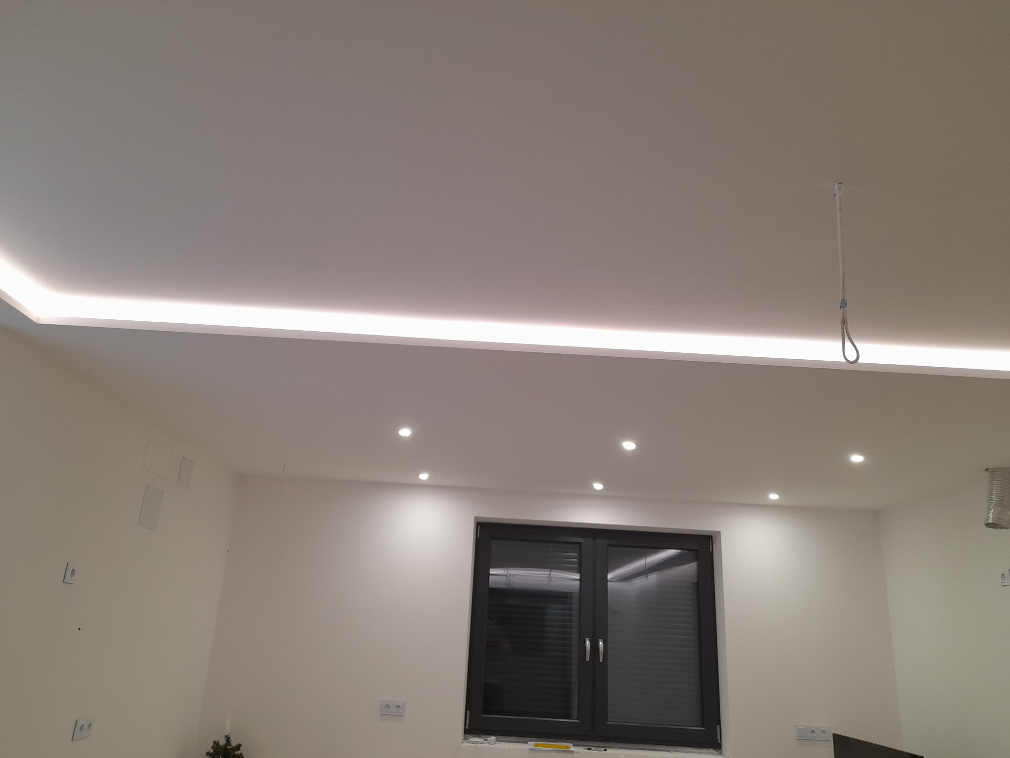 Białe wnętrze z podwieszonym sufitem z oświetleniem LED, widoczne punkty świetlne, okno w czarnej ramie i wystające kable ze stropu.