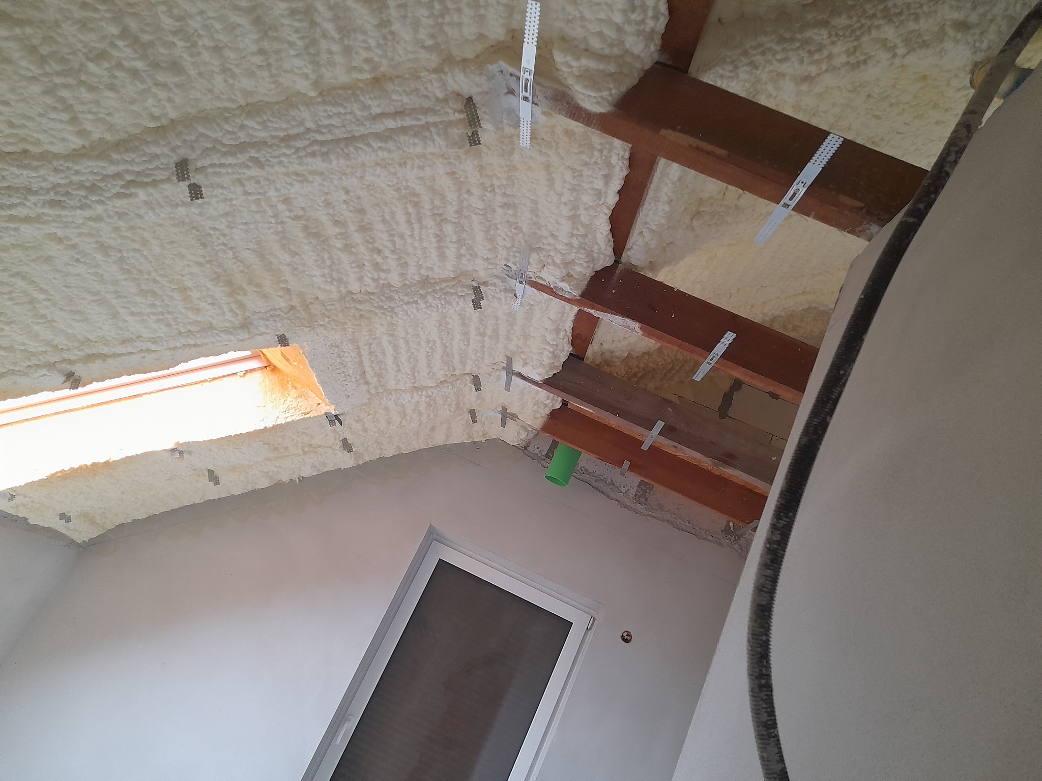 Poddasze ocieplone pianą poliuretanową, widoczne drewniane krokwie i metalowe elementy montażowe, okno dachowe.