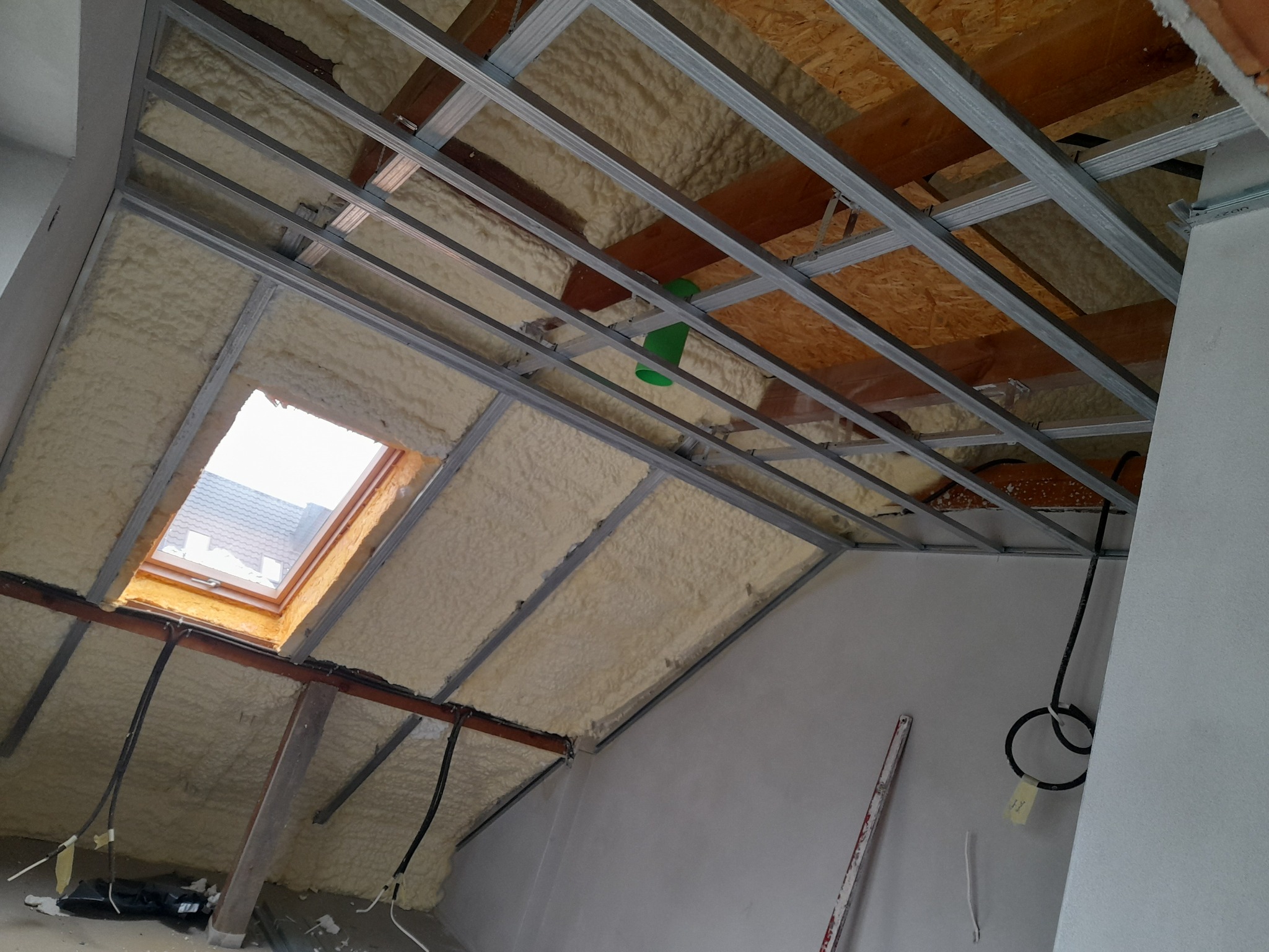 Skośny sufit poddasza z oknem dachowym, ocieplony pianką poliuretanową, z widoczną metalową konstrukcją pod zabudowę.