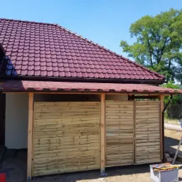 Zadaszone miejsce na śmietnik z drewnianymi przesuwnymi panelami, widoczny fragment dachu z czerwoną dachówką i rynną oraz narzędzia pracy.