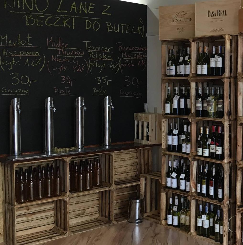 Ekspozycja win lanych z beczki do butelek w drewnianych skrzynkach, z tablicą informacyjną o gatunkach, cenach i pochodzeniu win.