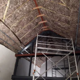 Ocieplenie poddasza wełną mineralną widoczne spod rusztowania, drewniane i metalowe elementy konstrukcyjne dachu, surowy stan wykończenia.