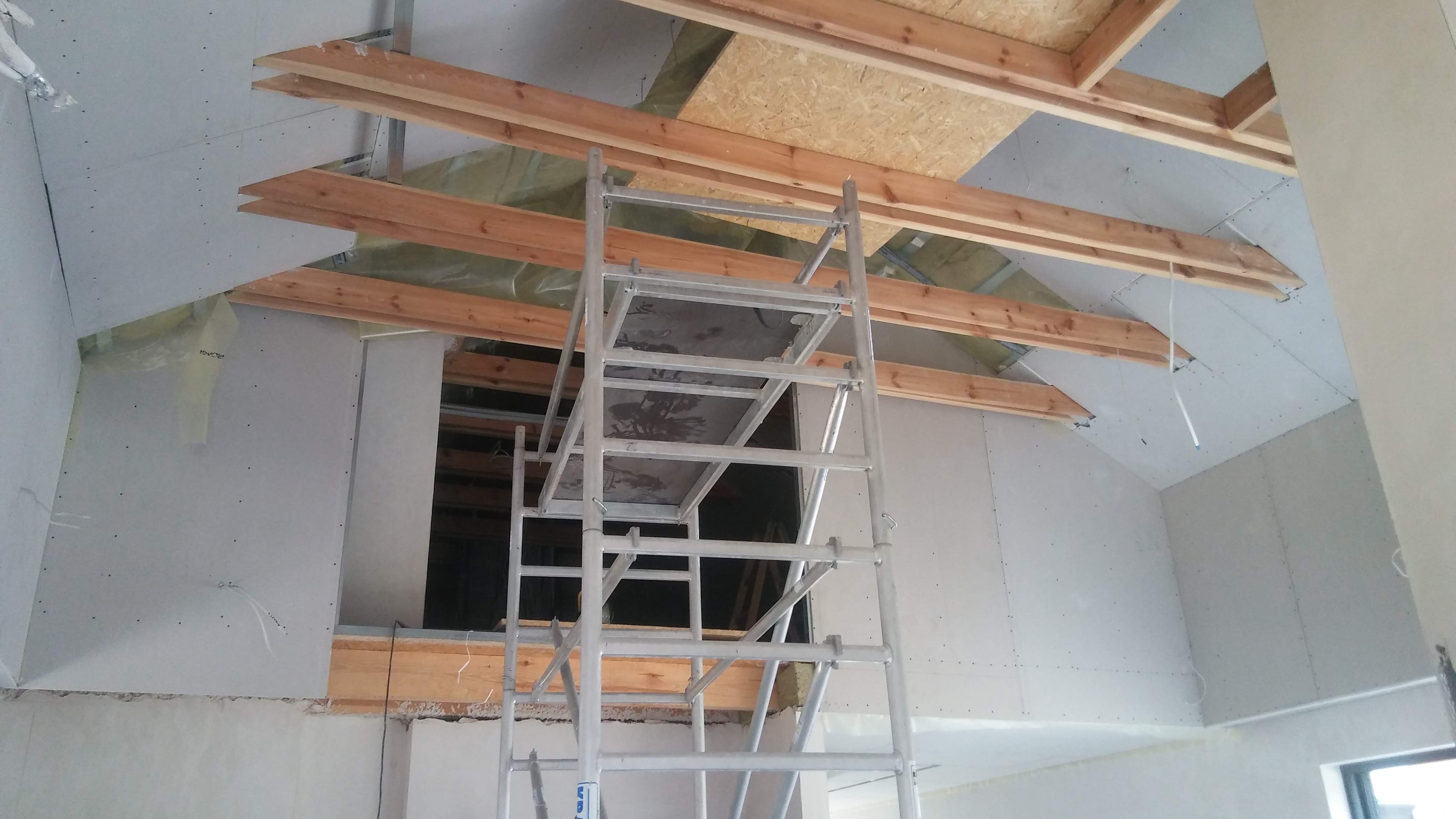 Stelaż rusztowania na tle poddasza w trakcie prac wykończeniowych, widoczne drewniane belki, płyty kartonowo-gipsowe oraz izolacja.