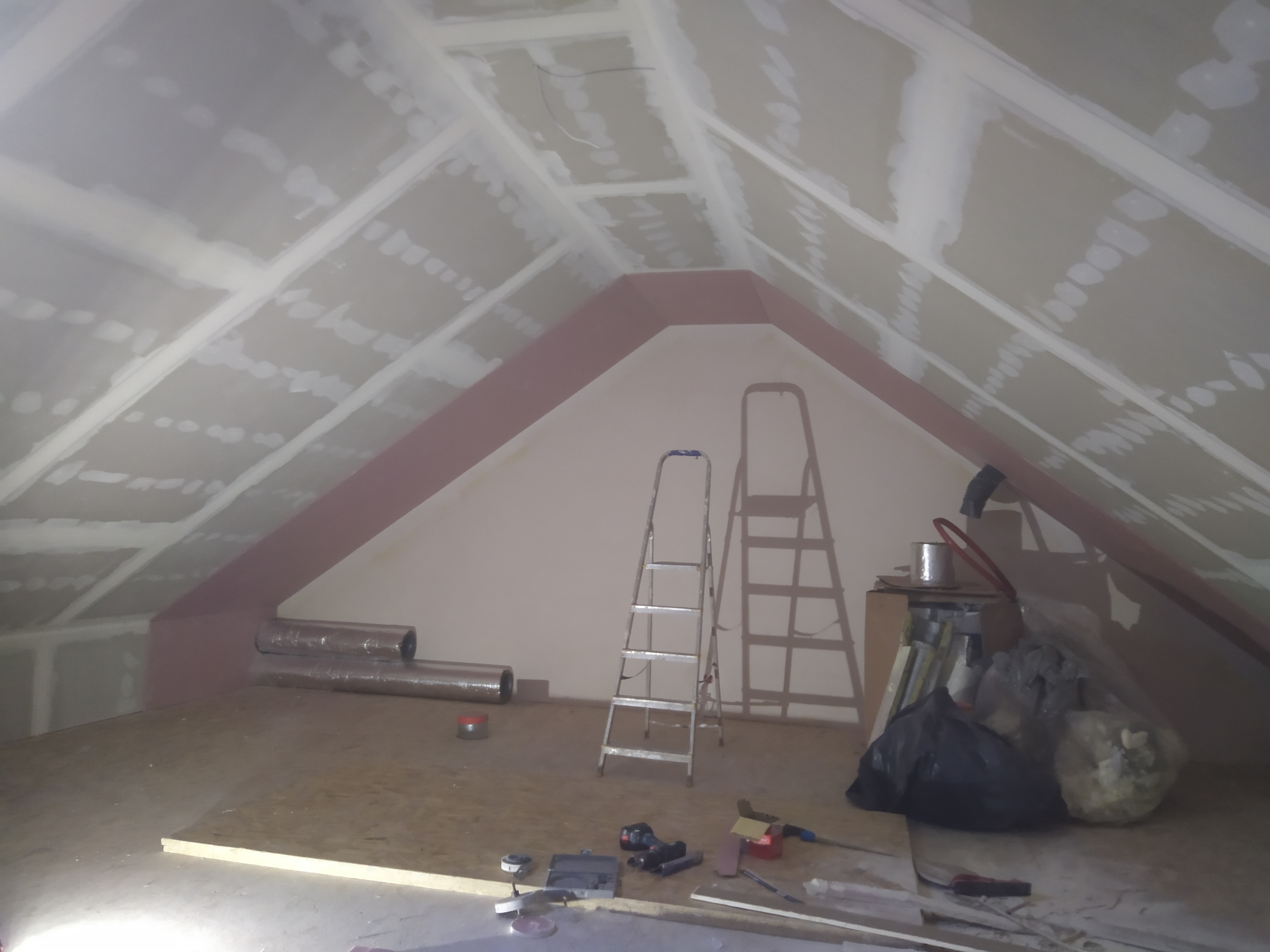 Poddasze w trakcie wykańczania płytami kartonowo-gipsowymi, widoczne profile, szpachlowane łączenia, dwie drabiny, rolki izolacji i narzędzia na podłodze.