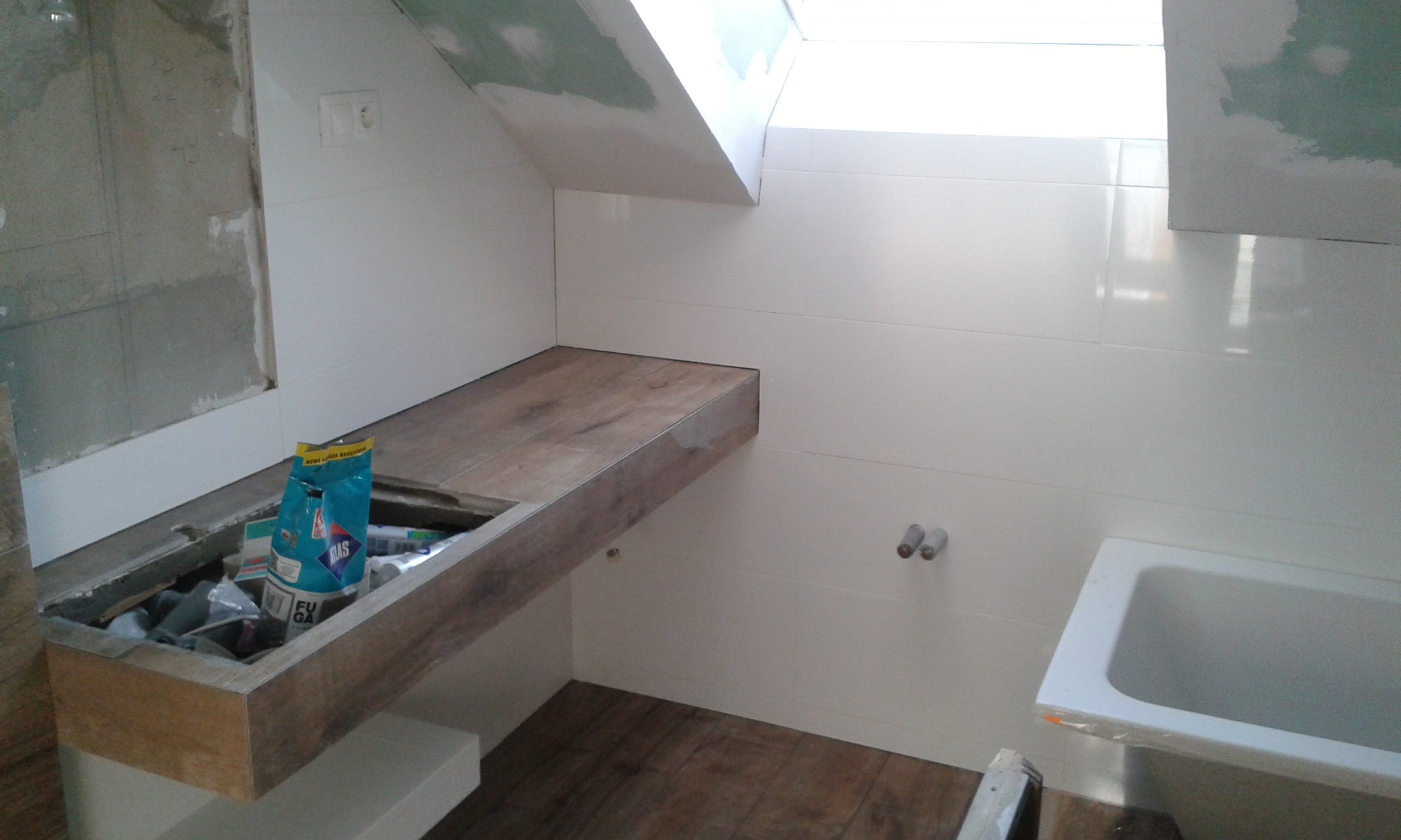 Łazienka w trakcie remontu na poddaszu: białe płytki ścienne, drewnopodobna zabudowa pod umywalkę z otwartą przestrzenią, widoczna wanna i materiały budowlane.