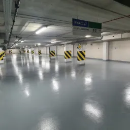 Szara, błyszcząca posadzka w garażu podziemnym z kolumnami oznaczonymi czarno-żółtymi pasami, widoczne znaki informacyjne i oświetlenie sufitowe.