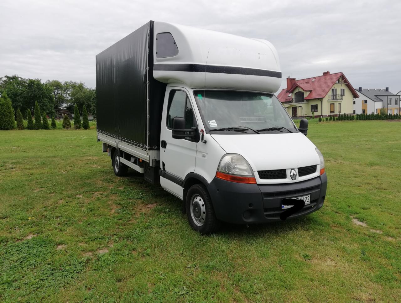 Biały samochód dostawczy Renault Master z czarną plandeką na tle zielonego trawnika i zabudowań mieszkalnych.
