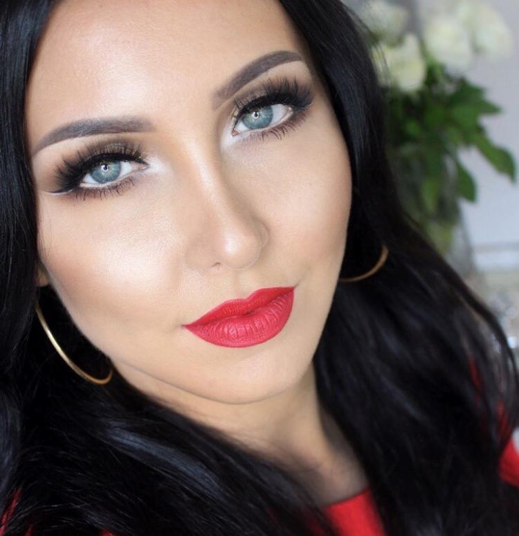 Portret kobiety z intensywnym makijażem: czerwone matowe usta, mocno podkreślone oczy z białym eyelinerem, długie sztuczne rzęsy i wykonturowana twarz. W tle widoczne białe kwiaty.