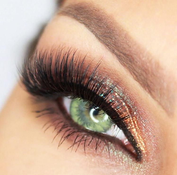 Zbliżenie na oko z zieloną tęczówką, podkreślone błyszczącym, miedziano-złotym cieniem i długimi, gęstymi rzęsami.