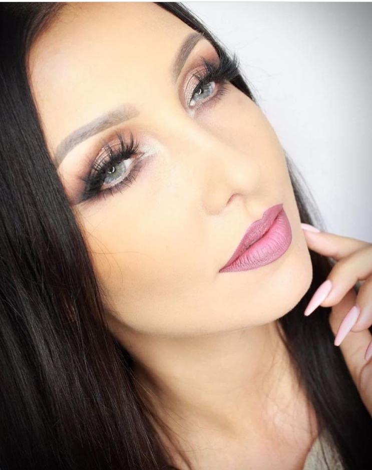 Portret kobiety z intensywnym makijażem oczu, sztucznymi rzęsami i matową pomadką w odcieniu różu, dłoń z jasnoróżowymi paznokciami dotyka policzka.