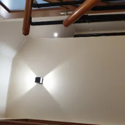 Nowoczesne wnętrze z beżowymi ścianami, drewnianą konstrukcją sufitową i minimalistycznym oświetleniem ściennym w kształcie sześcianu, rzucającym promienie światła w górę i w dół.