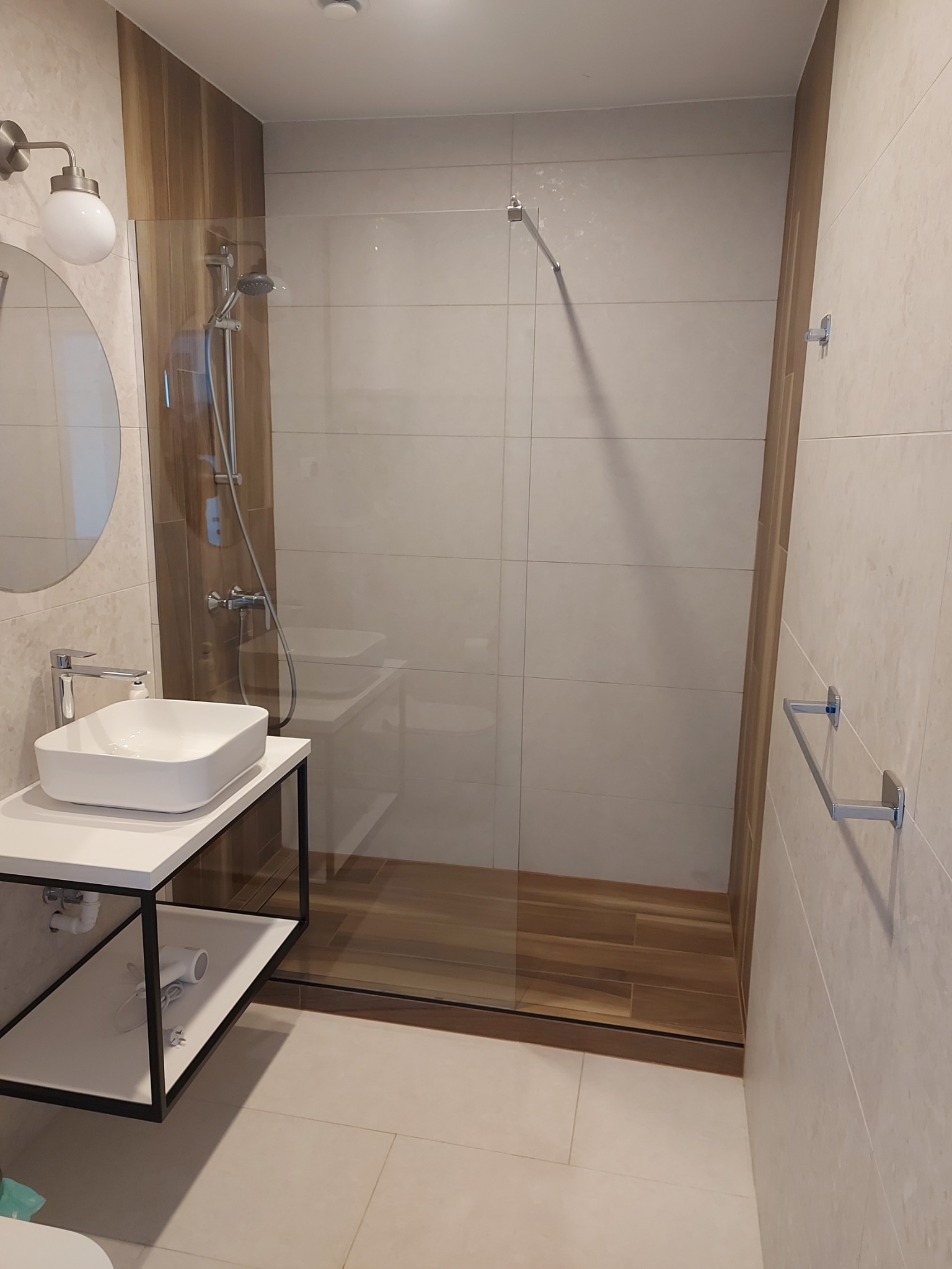 Nowoczesna łazienka po remoncie: kabina walk-in z drewnopodobną ścianą, umywalka nablatowa na czarnym stelażu, jasne płytki i oświetlenie.