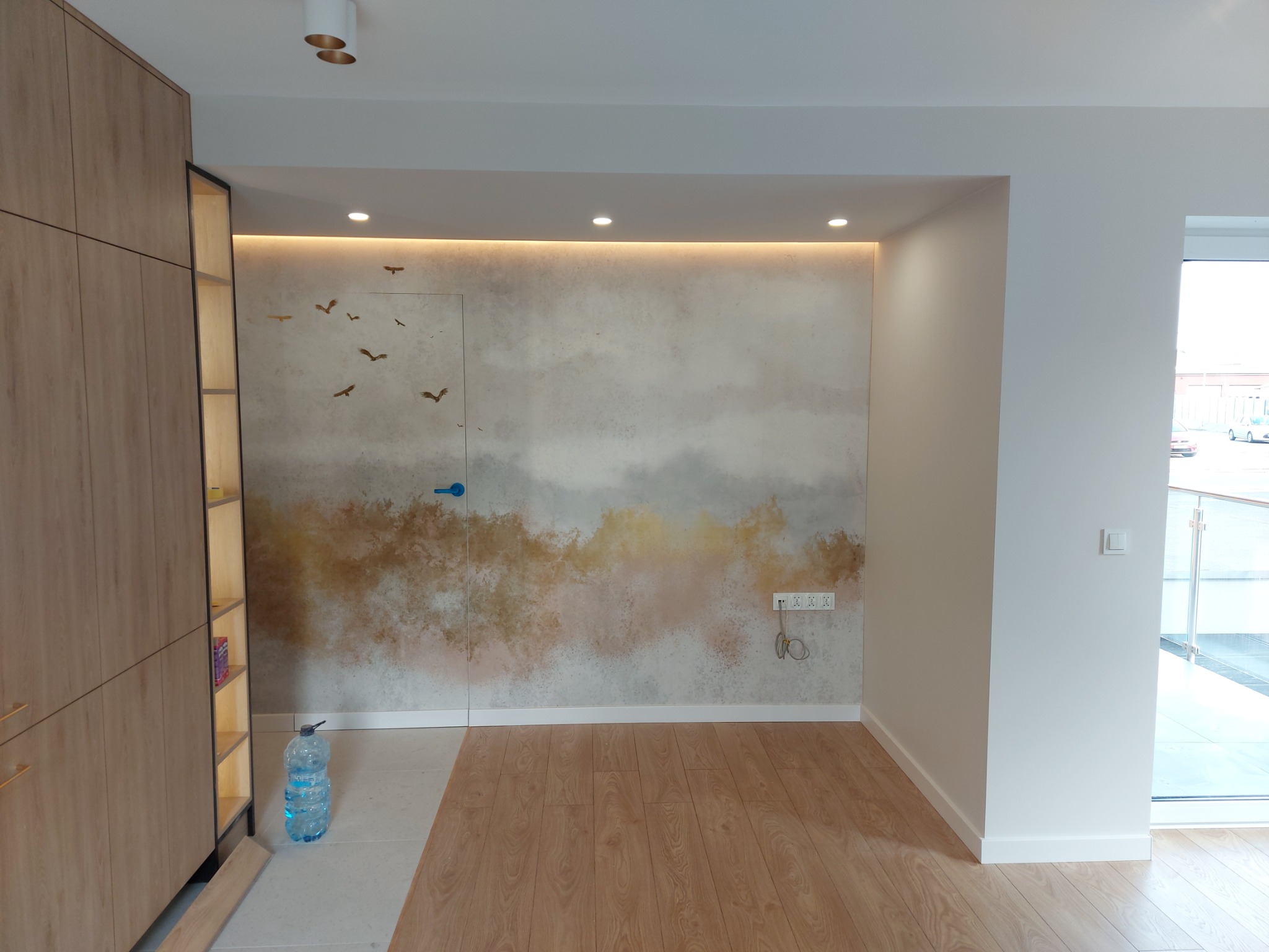 Nowoczesne wnętrze: drewniane panele podłogowe, ściana z motywem ptaków i ukryte drzwi z niebieską klamką. Minimalistyczny design z subtelnym oświetleniem LED.