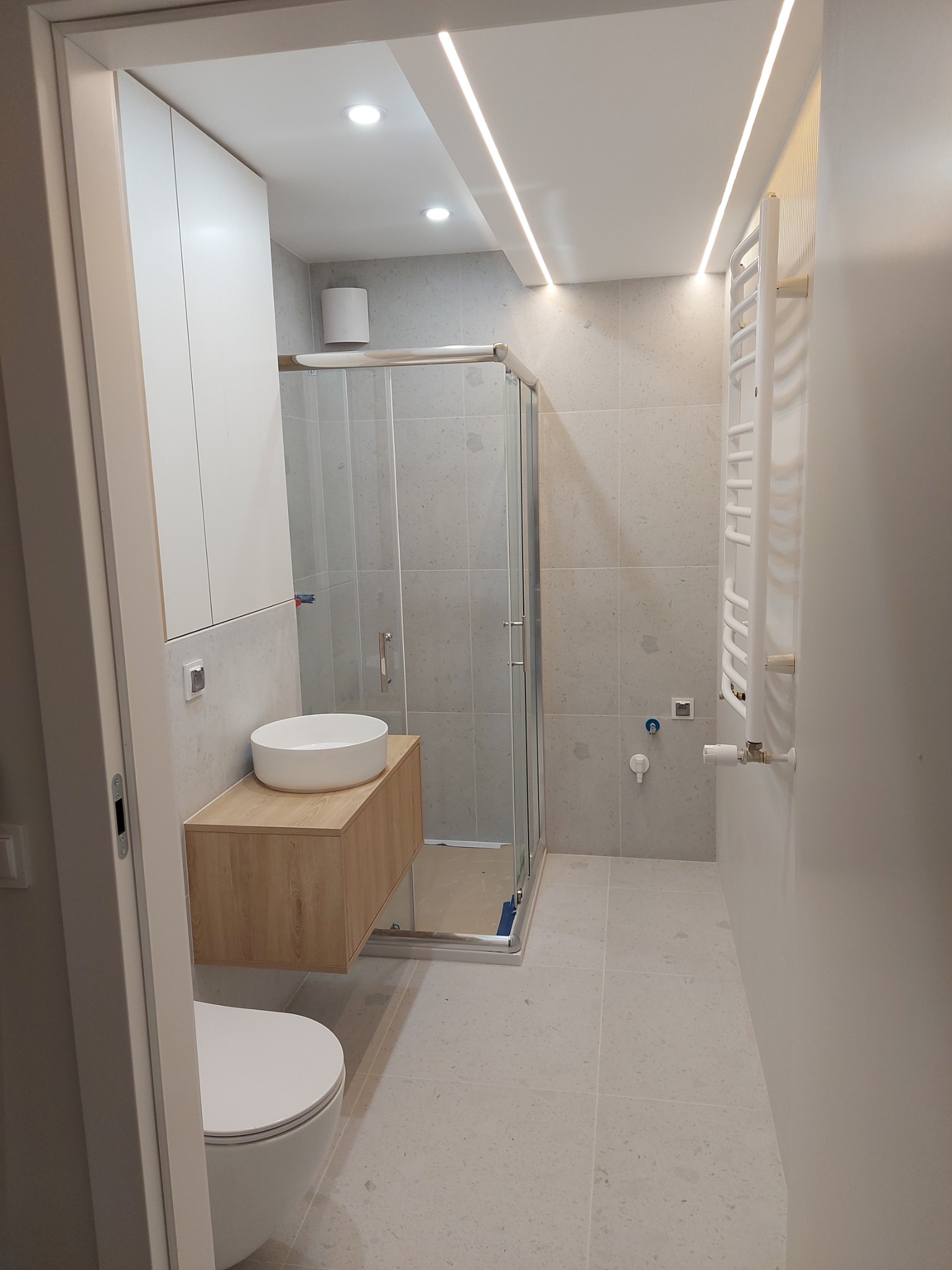 Nowoczesna łazienka w jasnych barwach z kabiną prysznicową, umywalką na drewnianej szafce i grzejnikiem drabinkowym. Oświetlenie LED podkreśla minimalistyczny design.